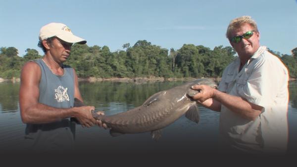 Two men hold up a giant catfish with the river in background