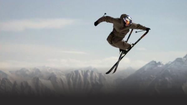 Skier looks over his shoulder mid-turn, skis crossed, with mountain peaks in background