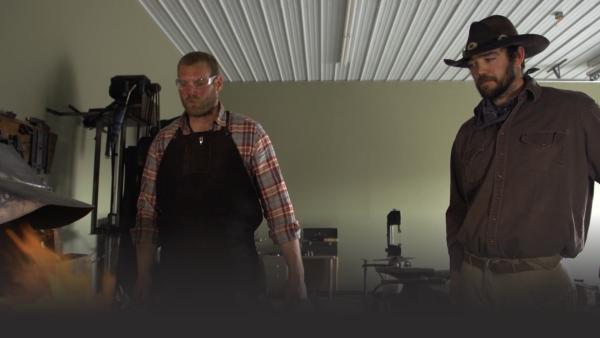 Two men stand over a fire in a blacksmith shop.