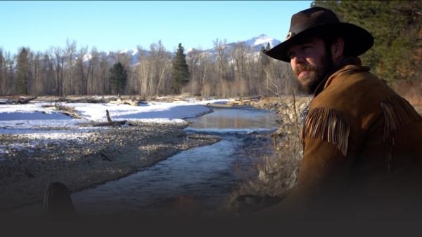 A man sits next to a fire by a river.