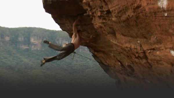 Man climbing has jumps up the cliff to grab above him, both legs swinging out